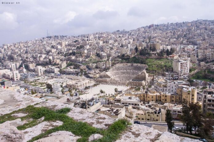 fotografia colori, panorama diurno città Amman, resti archeologici primo piano, sfondo edifici moderni su collina, colori chiari bianco grigio azzurro