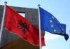 Fotografia colori, esterno, a sinistra bandiera albanese aquila nera due teste in campo rosso, a destra bandiera europea 12 stelle gialle in cerchio su campo blu