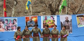fotografia collri, esterno, 6 donne curde combattenti unità delle donne libere, vestite di verde, sfondo telo azzurro con fotografie, bandiere e alberi