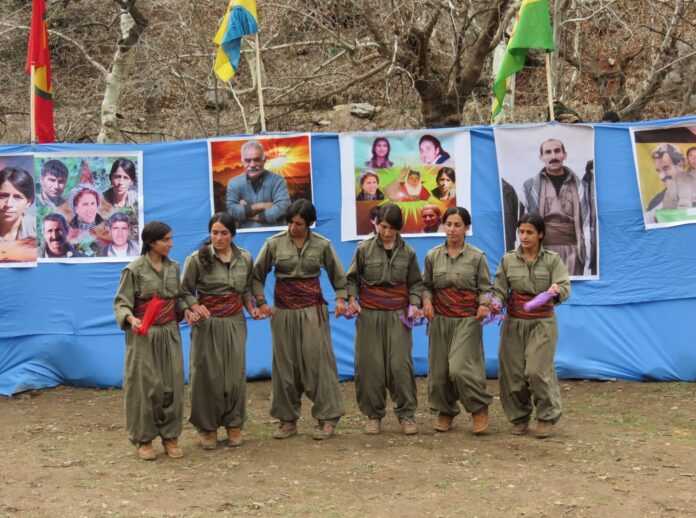 fotografia collri, esterno, 6 donne curde combattenti unità delle donne libere, vestite di verde, sfondo telo azzurro con fotografie, bandiere e alberi