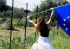 fotografia, diurno esterno, opera di Marilena Vita, ragazza di spalle agita bandiera europea accanto a recinzione metallica, sfondo prato e alberi