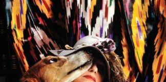 fotografia colori, la poetessa Evelina Schatz seduta con in braccio un cane marrone, vestita con scialle bianco e cappello con fiori e piume, sfondo tendaggio fantasia multicolore