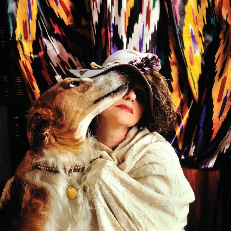 fotografia colori, la poetessa Evelina Schatz seduta con in braccio un cane marrone, vestita con scialle bianco e cappello con fiori e piume, sfondo tendaggio fantasia multicolore