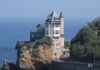 fotografia, colori, esterno, castello bianco su sperone roccioso in riva al mare, alberi a destra, sfondo mare e cielo azzurri