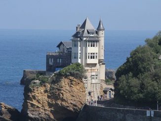 fotografia, colori, esterno, castello bianco su sperone roccioso in riva al mare, alberi a destra, sfondo mare e cielo azzurri
