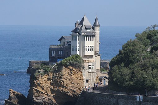 fotografia, colori, esterno, castello bianco su sperone roccioso in riva al mare, alberi a destra, sfondo mare e cielo azzurri