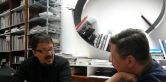 Fotografia colori interno, due uomini in conversazione, seduti alla scrivania, entrambi occhiali e vestiti scuro, sfondo libreria