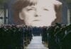 Angela Merkel versione "1984" di Gerge Orwell, la sua faccia è proiettata sullo schermo davanti ai suoi sudditi.
