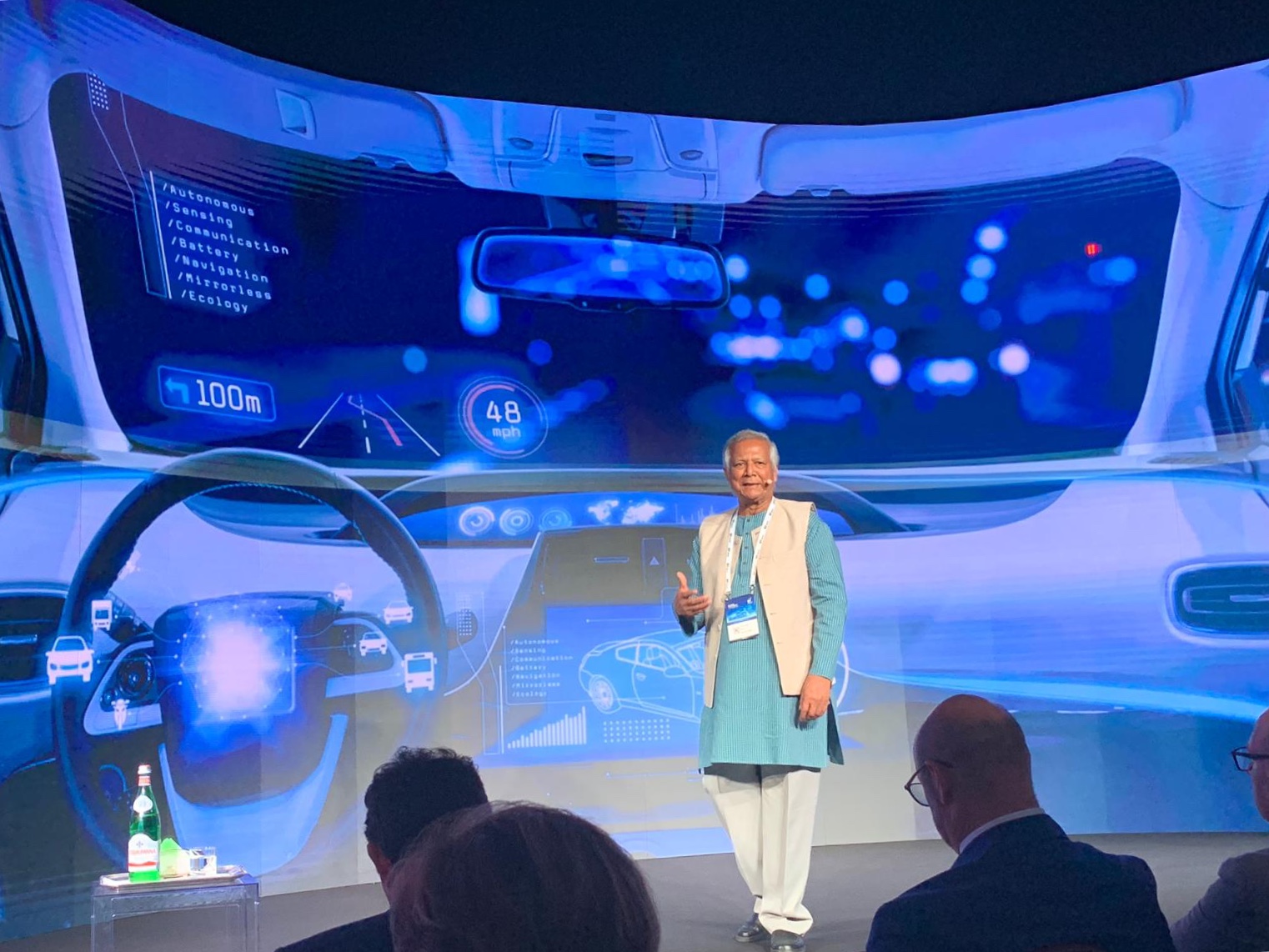 Fotografia dell'economista e professore bengalese Mohammad Yunus mentre parla all'uditorio di EY Digital Summit 2019. Yunus indossa abiti tradizionali del proprio paese, pantaloni lunghi chiari sotto una tunica celeste e un gilet chiaro.
