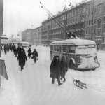 Una strada di Leningrado innevata, con persone che camminano, un filobus che transita sulla carreggiata colma di neve