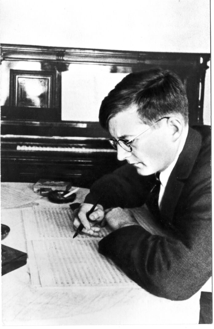 Fotografia del compositore Dmítrij Dmítrievič Šostakóvič seduto alla sia scrivania, chino sul pentagramma. Sullo sfondo un pianoforte verticale