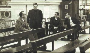 fotografia, bianco e nero, interno chiesa, 3 uomini in giacca e cravatta seduti sulle panche della chiesa, prete al centro in piedi