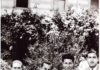 Black-and-white photography with 4 men in suits sitting on a bench, with shrubs on the background