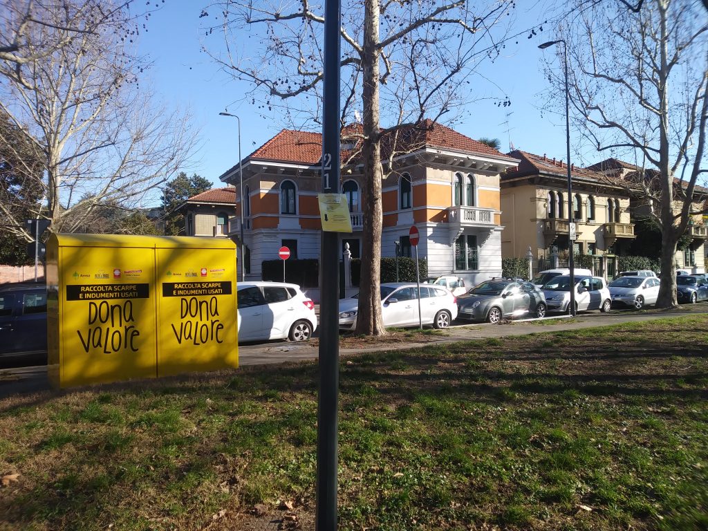 Una foto orizzontale che mostra un'aiuola nella periferia milanese. al centro un palo sul quale è appeso un piccolo foglio giallo plastificato. Sulla sinistra sono visibili due punti di raccolta per scarpe e indumenti usati. Una strada separa lo spazio verde da alcune case a due piani
