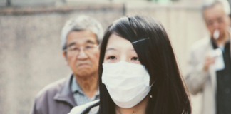 Una donna dai tratti asiatici indossa una mascherina bianca che copre il naso e la bocca. Dietro si intravedono due uomini