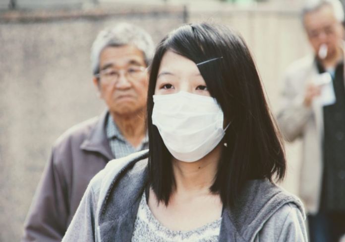 Una donna dai tratti asiatici indossa una mascherina bianca che copre il naso e la bocca. Dietro si intravedono due uomini