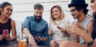 Un allegro gruppo di amici, composto da tre ragazze e due ragazzi, gioca a carte davanti ad un tavolo sul quale sono appoggiati anche un bicchiere con una bibita color arancio e un telefono cellulare.