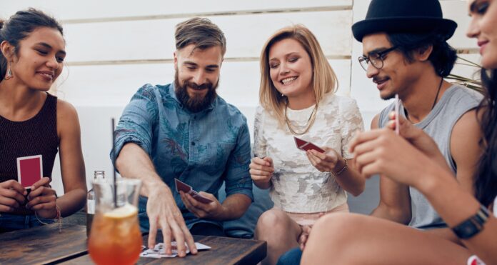 Un allegro gruppo di amici, composto da tre ragazze e due ragazzi, gioca a carte davanti ad un tavolo sul quale sono appoggiati anche un bicchiere con una bibita color arancio e un telefono cellulare.