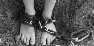 Una foto in bianco e nero mostra i piedi nudi di una persona poggiati sulla terra e immobilizzati da una catena