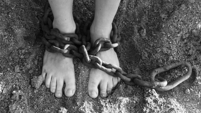Una foto in bianco e nero mostra i piedi nudi di una persona poggiati sulla terra e immobilizzati da una catena