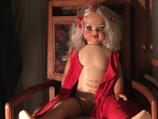 Bambola con capelli biondi fermati all'altezza delle tempie da due fermagli rossi, indossa un abito rosso completamente aperto sul fronte. La bambola è seduta su una sedia di legno appoggiata ad una parete lignea.