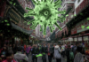 L'immagine mostra una affollata via cinese e sospesa sulle teste delle metaforicamente incombe una grande struttura verde che riproduce la struttura di un virus-