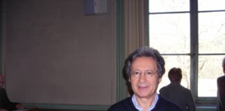 fotografia, colori, interno, ritratto di Angelo Gaccione scrittore italiano e poeta, in aula presso università di Ginevra, uomo brizzolato, occhiali, maglione blu e camicia azzurra