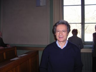 fotografia, colori, interno, ritratto di Angelo Gaccione scrittore italiano e poeta, in aula presso università di Ginevra, uomo brizzolato, occhiali, maglione blu e camicia azzurra