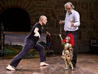 A man with grey hair manipulates a puppet withstanding a pointed object. Another man with naked feet opens his harm in front of the puppet. In the surrounding environment there are a chair and a painting
