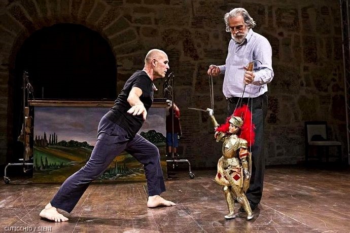Danza e marionetta Un uomo con barba e capelli bianchi regge i fili di una marionetta. La marionetta ha in mano un oggetto appuntito e di fronte a lei un uomo a piedi nudi spalanca le braccia. Nell'ambiente dietro di loro si vedono un quadro a sinistra e una sedia a destra