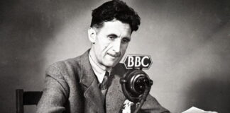 Fotografia in bianco e nero dello scrittore britannico George Orwell, seduto alla scrivania di fronte a un microfono della BBC.