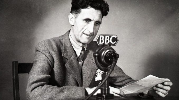 Fotografia in bianco e nero dello scrittore britannico George Orwell, seduto alla scrivania di fronte a un microfono della BBC.