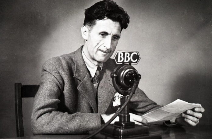 Fotografia in bianco e nero dello scrittore britannico George Orwell, seduto alla scrivania di fronte a un microfono della BBC.