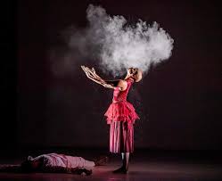 Una ballerina di profilo, in piedi, braccia aperte invocanti il cielo, capo piegato all'indietro, abito di scena rosso. Ai suoi piedi giace un uomo.