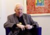 Il critico d'arte e poeta Edward Lucie-Smith seduto su una poltrona, sullo sfondo un'opera d'arte con colori accesi.