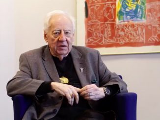 Il critico d'arte e poeta Edward Lucie-Smith seduto su una poltrona, sullo sfondo un'opera d'arte con colori accesi.