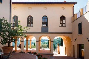Cortile interno di un edificio; al piano terra un porticato lascia intravedere delle persone sedute e sullo sfondo un paesaggio. In primo piano a sinistra c'è una pianta dentro a un vaso