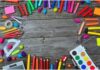 Matite colorate, pastelli a cera, tavolozza con colori all'acqua, un compasso, una calcolatrice, un temperino, un goniometro: strumenti di lavoro per uno scolaro, sparsi su un tavolo in legno.