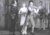 The picture is a black and white photography that represent a scene from the ballet "La Strada" by and with Mario Pistoni. From Left to right Aldo Santambrogio (as Zampnò, dressed in black), Carla Fracci (as Gelsomina, dressed in white), Mario Pistoni (as the foul, dressed in strips and playing a violin). On the right, in front of a tarp that covers the backdrop, there are two ballet dancers dressed with sequin costumes