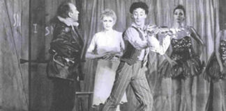 The picture is a black and white photography that represent a scene from the ballet "La Strada" by and with Mario Pistoni. From Left to right Aldo Santambrogio (as Zampnò, dressed in black), Carla Fracci (as Gelsomina, dressed in white), Mario Pistoni (as the foul, dressed in strips and playing a violin). On the right, in front of a tarp that covers the backdrop, there are two ballet dancers dressed with sequin costumes