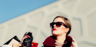 donna con capelli legati, occhiali da sole, rossetto sulle labbra e sguardo rivolto lateralmente regge nella mano destra una serie di buste di colori diversi. La donna indossa capi d'abbigliamento invernali: sciarpa, cappotto, guanti in pelle