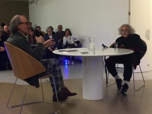 Photograph of the presentation of the autobiography of Nanda Vigo moderated by Carmelo Strano. The two are in a room, around a table, with an audience listening.