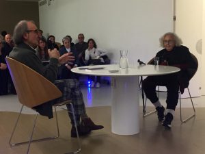 Fotografia della presentazione dell'autobiografia di Nanda Vigo moderata da Carmelo Strano. I due sono in una sala, intorno a un tavolo, con pubblico che ascolta.