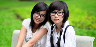 Due giovani ragazze dai tratti orientali, entrambe con occhiali da vista, posano sedute in un'area verde. Una di loro guarda verso l'osservatore, l'altra rivolge lo sguardo lateralmente