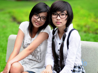 Due giovani ragazze dai tratti orientali, entrambe con occhiali da vista, posano sedute in un'area verde. Una di loro guarda verso l'osservatore, l'altra rivolge lo sguardo lateralmente