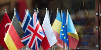 Su un tavolo di legno, di fronte ad una superficie riflettente sono disposte dodici bandierine. quelle riconoscibili sono -da sinsitra verso destra- Turchia, Germania, Grecia, Regno Unito, Polonia, Moldavia, Unione Europea e Romania