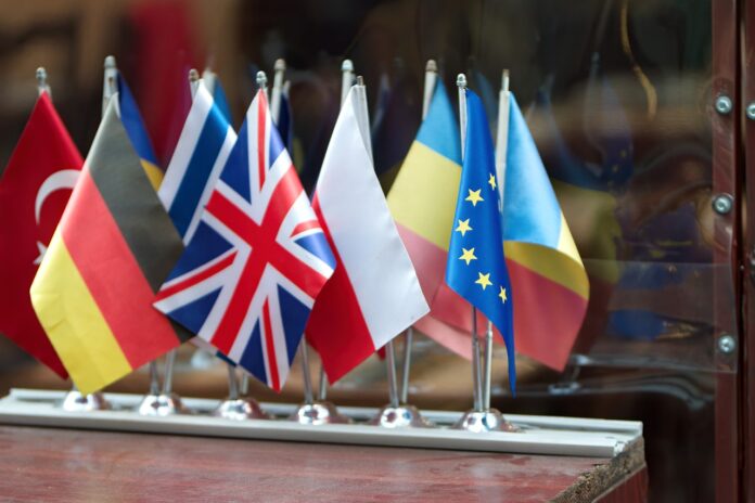 Su un tavolo di legno, di fronte ad una superficie riflettente sono disposte dodici bandierine. quelle riconoscibili sono -da sinsitra verso destra- Turchia, Germania, Grecia, Regno Unito, Polonia, Moldavia, Unione Europea e Romania