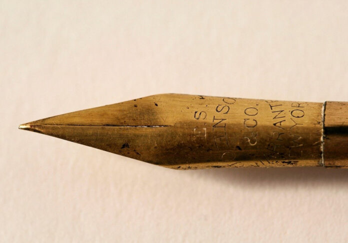 Punta di una penna color oro rivolta verso sinistra e con lettere incise sopra