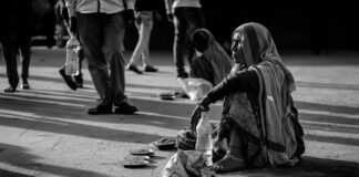 The picture is a black and white photography. The image shows an indian road with several people walking around. In the foreground, a barefoot woman, dressed in poor clothes is sitting on the ground, with a pair of sandal, a bottle of water and some little bowl with seeds inside. Another sitting woman can be seen in the background. Both the women are overlooked by the passers-by