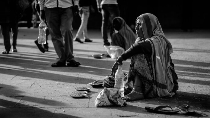 L'immagine è una foto in bianco e nero che mostra una strada attraversata da diverse persone riprese dal basso. In primo piano una donna vestita con abiti tradizionali indiani è seduta per terra, con un paio di sandali, una bottiglia d'acqua e quattro ciotole piene di semi. In secondo piano è visibile un'altra donna, anch'essa seduta per terra ed anch'essa ignorata dai passanti